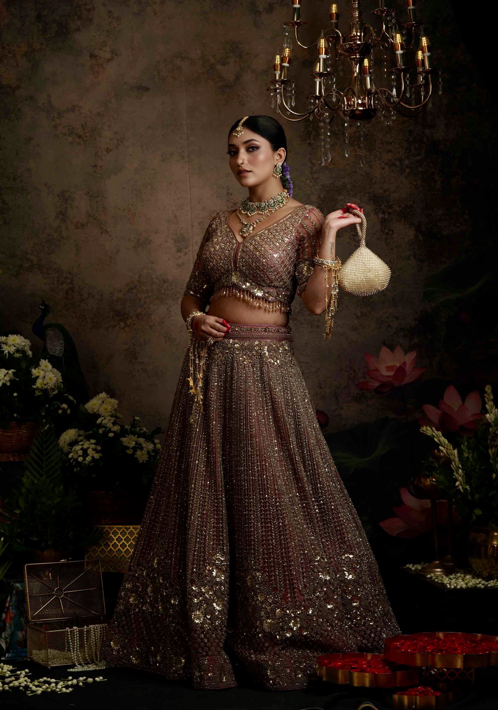 Bride in a bronze lehenga holding a gold evening bag beneath a chandelier