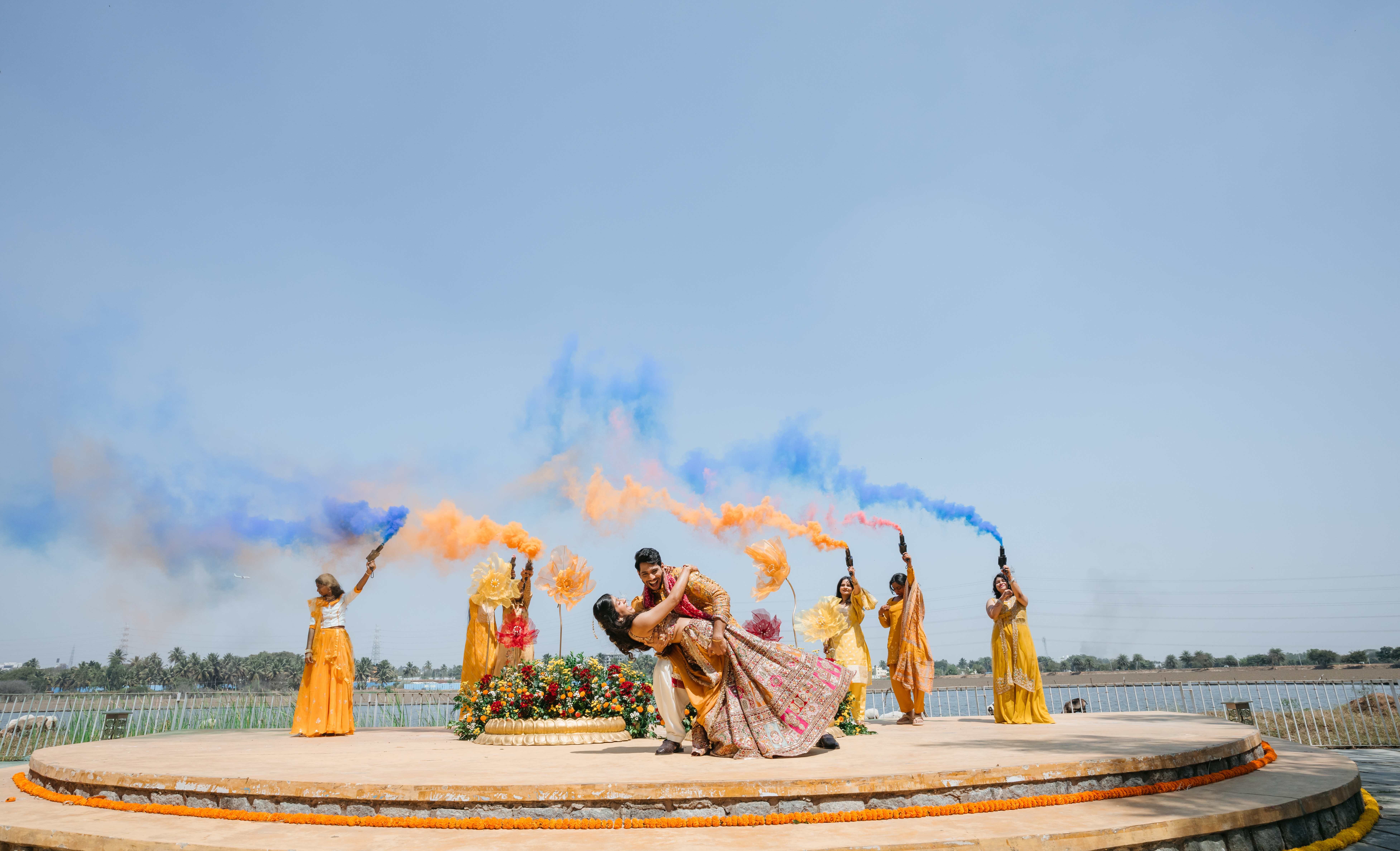 Bride and groom celebrating beneath blue and orange ceremonial smoke with friends around them