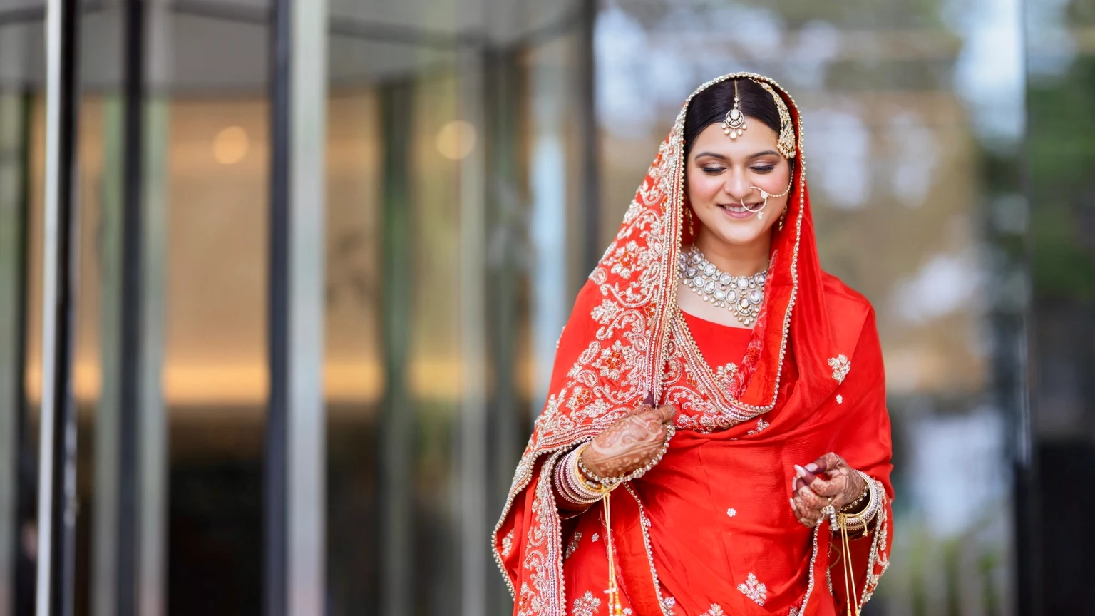 Bride in red wedding attire standing beside a glass exterior in soft daylight.